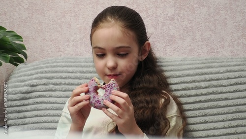 A portrait of a beautiful little girl holding a dessert—a donut covered in icing. A sweet expression on the girl's face as she enjoys a sweet donut. Diet for weight loss. Eating fast food and unhealth