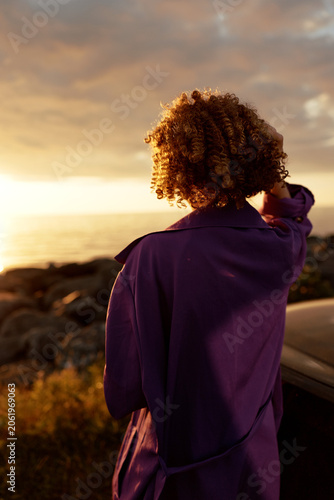 Foto A back view of a woman in a purple coat outdoors at sunset, a gentle breeze lifting curls, with a thoughtful silhouette against the horizon and warm light