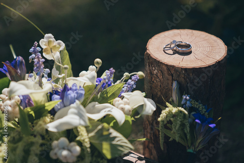 Wedding rings displayed on rustic wood slice beside bridal bouquet at outdoor wedding ceremony, Close up of wedding rings on wooden stand with bridal bouquet at outdoor wedding venue