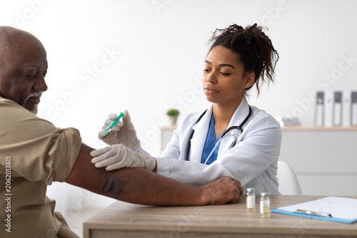 Tableau sur toile Focused Female Doctor Administering Vaccine To Elderly Black Male Patient In Modern Clinic, Ensuring Proper Care And Support During Medical Appointment