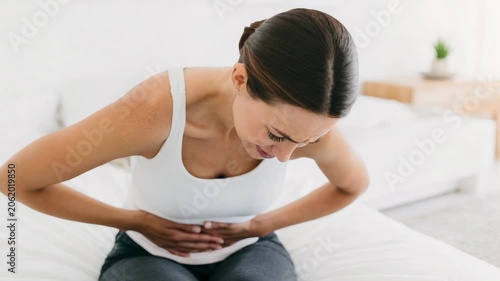 Woman sitting on bed holding abdomen in pain, wearing white tank top and grey leggings, indicating digestive issues, stomach cramps, or menstrual discomfort.