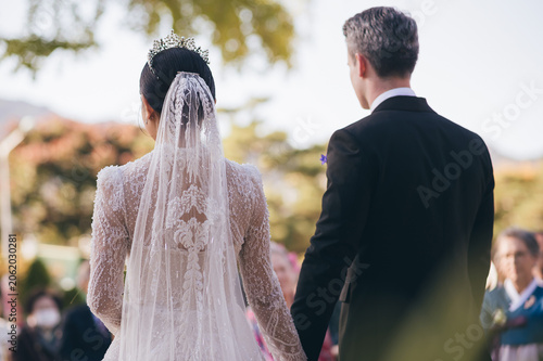 Back view of elegant intercultural bride and groom holding hands at outdoor wedding ceremony in South Korea