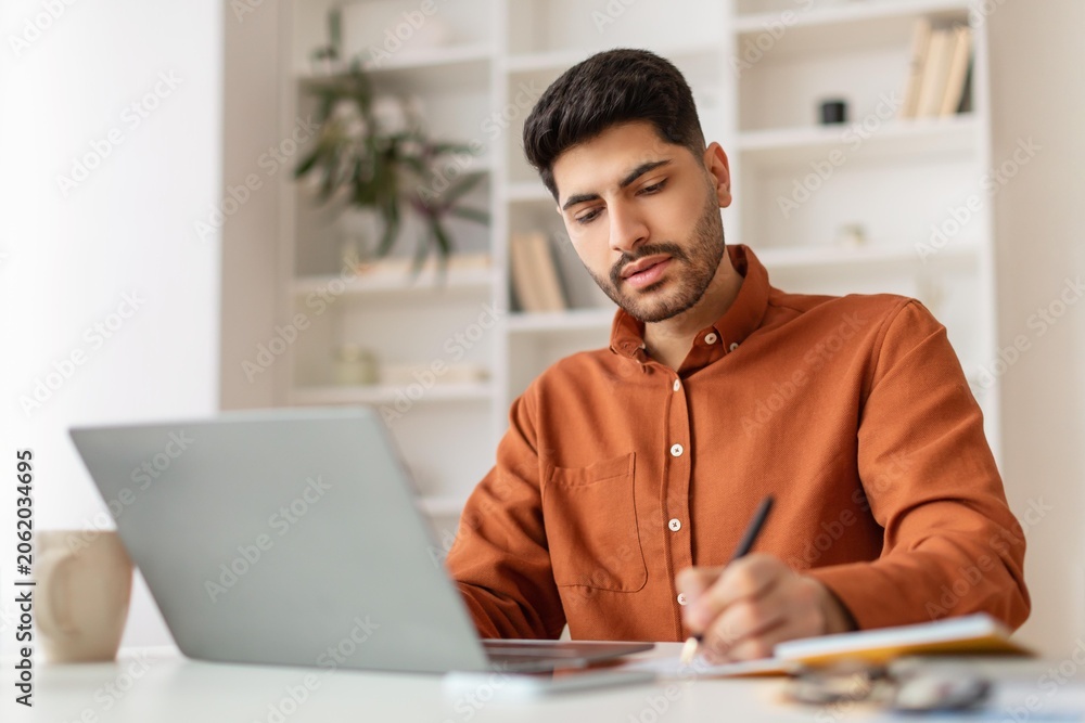 Fototapeta premium Focused Young Hispanic Man Working Diligently On Laptop While Taking Notes At Home Office, Surrounded By Shelves And Plants, Concentrated On Task. Professional Business Concept.