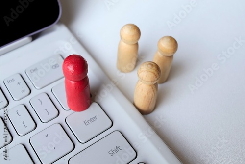 ​A red wooden peg doll standing on a laptop keyboard with natural figures in the background, concept of remote work, digital leadership, and online recruitment.