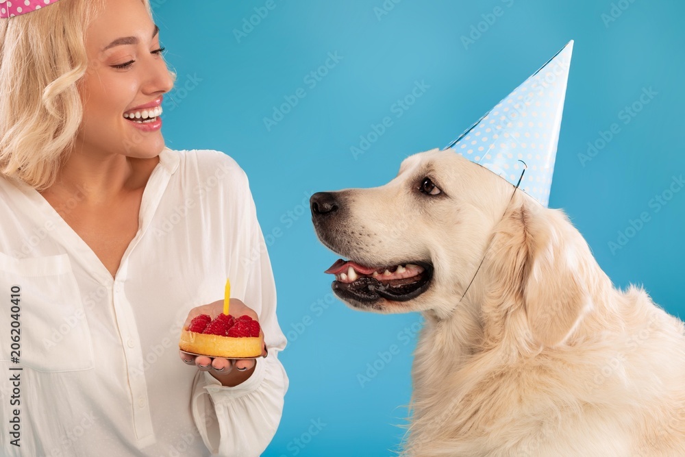 Fototapeta premium Smiling Young Caucasian Woman Celebrating Dog's Birthday By Holding Cake With Candle, Happy Atmosphere In Studio Setting, Joyful Moment With Pet. Family And Pet Celebration Concept.