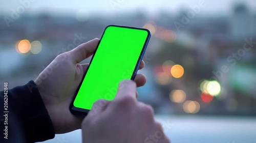 Close-Up of a Man's Hands Using a Smartphone with a Green Screen Against a Blurred Urban Evening Background