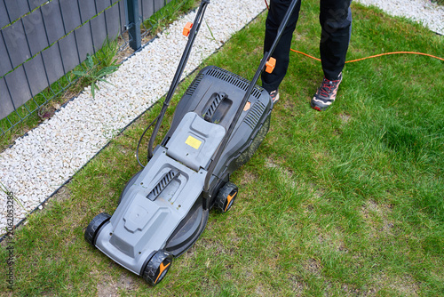 Lawn mower with power cord stands on backyard grass near fence and gravel border