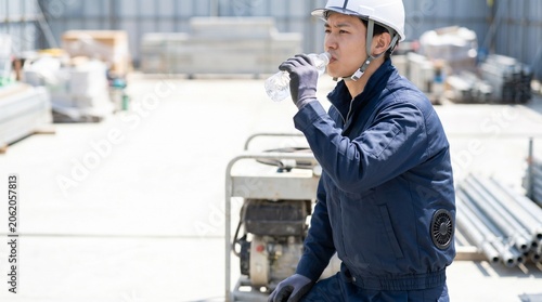 機材のある建設現場で空調服を着て水分補給による熱中症対策を行う20代30代の日本人男性の現場監督