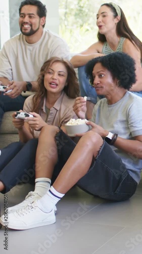 Vertical video: Playing friends leaning at home with gamepads reacting to game and sharing popcorn