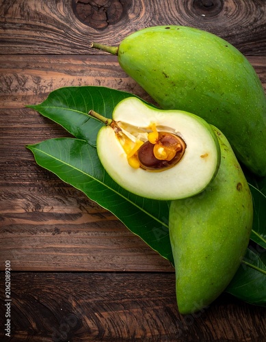 A halved green mango on leaves atop a wooden surface with two whole mangoes