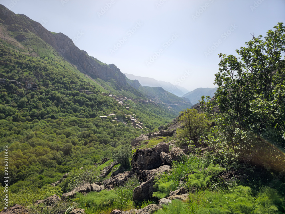 Obraz premium Deep mountain valley with rocky foreground, lush spring orchards, a distant hillside village, and layered peaks under bright sky. 📍 Hawraman Takht, Kurdistan, Iran 