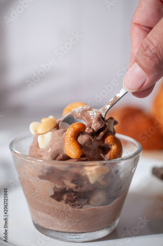 Chocolate ice cream inside a glass cup, with cashew nuts and white chocolate chips on top, illuminated by summer sunlight from a window, with a white wooden wall in the background. hand grabbing spoon