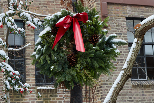snow covered christmas wreath decorated with pine cones and a red velvet bow hung on a pole outdoors in front of a brick building with holly branches on the side,
