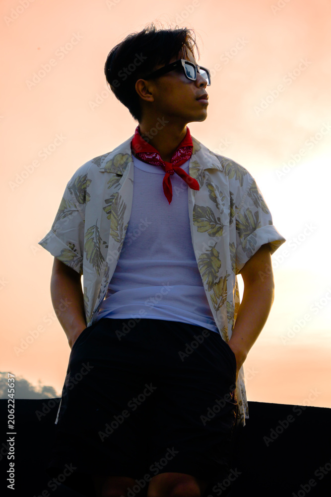 Fototapeta premium Stylish young Asian man in retro summer outfit posing outdoors against golden sunset sky background