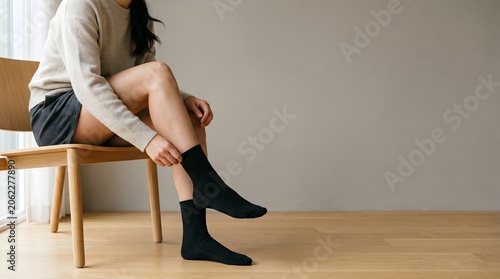 Young woman putting on comfortable black socks while sitting on a wooden chair at home