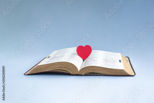 Red paper heart on top of open Holy Bible. Isolated on light blue background.