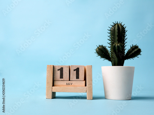 A wooden block calendar for July eleventh with a plastic cactus plant on the side. Simple and clean design on a soft blue background.