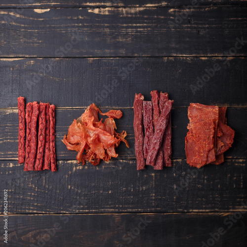 Dry salmon fish, chicken, red beef sticks and pork slices. Jerky meat on black wooden background. No brand, product presentation. Copy space, top view.