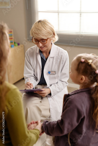 Vertical shot of Caucasian female pediatrician consulting child with mother, taking notes during checkup. Ideal for pediatric care, family health, medical services marketing