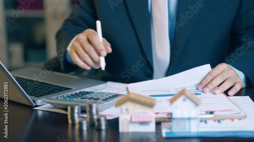 Businessman holding a pen, pointing at a house model and financial charts to review property contract details.