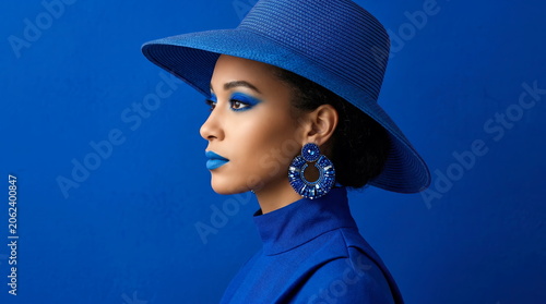 A young, beautiful, and sophisticated brunette woman posing against a blue background, wearing a hat, makeup, and clothing in the same shade of blue, striking a pose with her eyes open and expressing 
