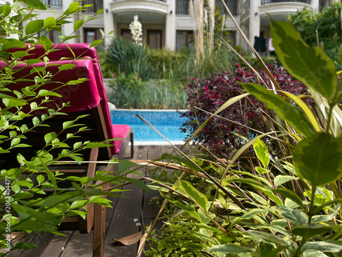 Horizontal close up shot of a luxury resort swimming pool seen through lush green garden plants and bushes with a pink sunbed chaise lounge on a wooden deck