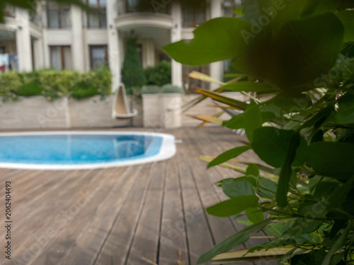 Horizontal photo of a luxury resort swimming pool with clear blue water on a wooden deck, viewed through lush green garden leaves with a hotel building in the blurred background