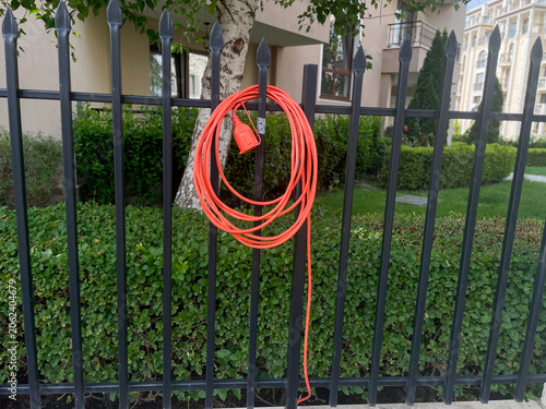 Horizontal photo of a bright orange heavy duty extension cord coiled and hanging on a black metal picket fence in front of a residential garden and building