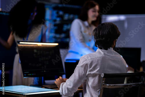 Red-haired female professional in a cream jacket holds documents and speaks to a Black female analyst beside a glowing monitor in a dark tech room.