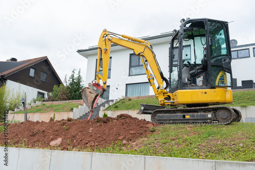 Compact excavator on slope near concrete wall. Residential garden terrace earthworks process