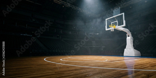 A basketball hoop stands on a wooden court with white lines in a darkened indoor arena with spotlights shining down.