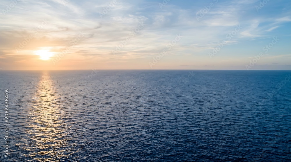 Obraz premium A serene aerial view of the vast blue ocean at sunset with golden light reflecting on the water surface under a soft, hazy, and colorful sky horizon