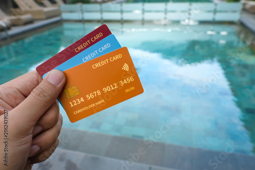 A hand holds several colorful credit cards in front of a swimming pool at a resort setting. Suitable for vacation payment, travel spending, cashless holiday, or credit card benefit