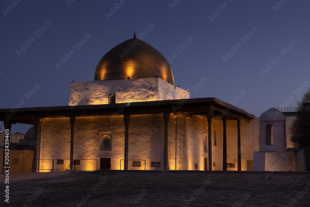 Naklejka premium Historic domed building illuminated at twilight inside Ichan Kala old town in Khiva, Uzbekistan. Warm uplighting highlights whitewashed walls and wooden columns against a deep blue evening sky.
