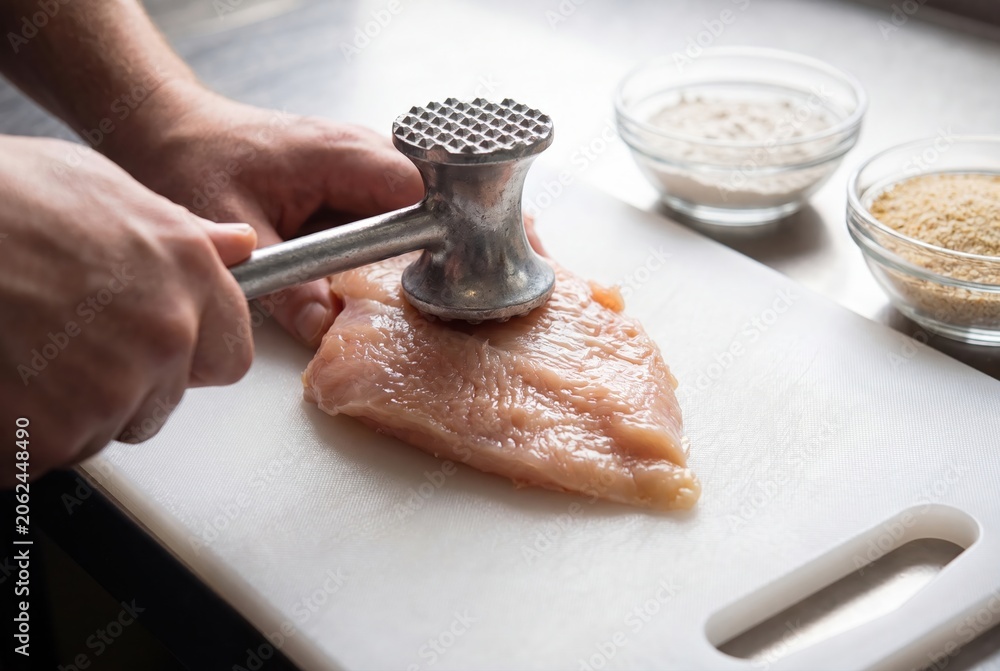 Fototapeta premium Chef tenderizing raw chicken breast with a metal meat mallet on a white cutting board, preparing for cooking.