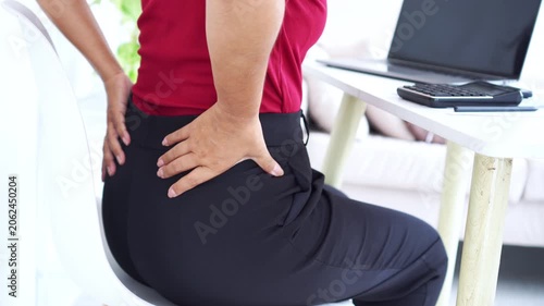 close-up of a woman sitting at her office desk holding her painful lower back, suffering from backache and office syndrome while working with a laptop and a calculator on a white tabl