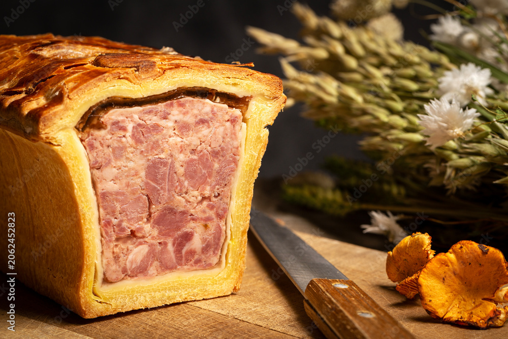 Obraz premium Pate en croute or pâté en croûte on a wooden cutting board, aside a knife and Chanterelle mushrooms on a dark gray background with dried flowers. Countryside vintage atmosphere.