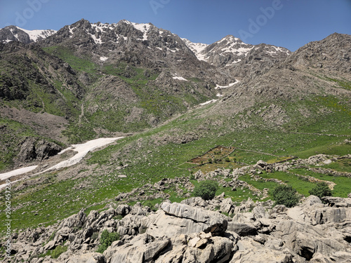 Fotografie Snow-capped highland peaks rise above a rugged green slope, with patches of snow lingering in the valley below and scattered shrubs across the spring landscape
