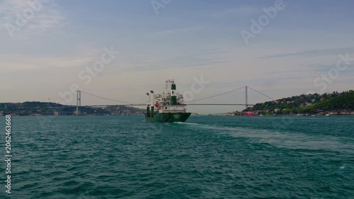 A large green cargo vessel cruising through the blue waters of the Bosphorus strait in Istanbul with a suspension bridge and hilly coastline in the background during daytime
