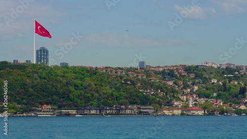 Large Turkish flag fluttering against a blue sky overlooking the green hills and residential buildings of the Istanbul coastline along the Bosphorus