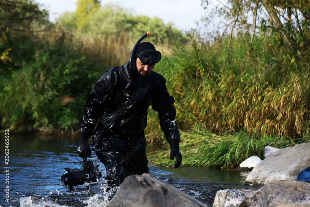 Naklejka premium Underwater Videographer in Black Wetsuit Navigating River Rocks Amidst Green Foliage