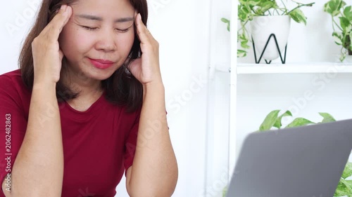 Asian woman in a red shirt touching her temples with closed eyes, feeling stressed, tired, or having a headache while working on a laptop at a bright home office desk, representing burnout