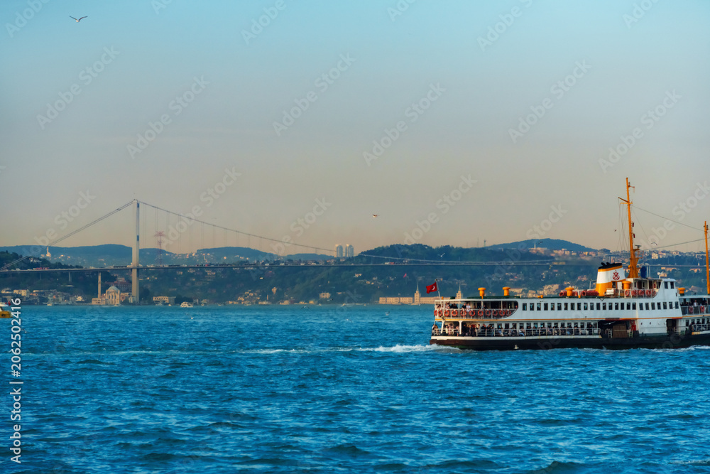 Fototapeta premium Traditional Passenger Ferry Boat Cruising on the Bosphorus Strait with the Iconic Bosphorus Bridge and City Skyline of Istanbul, Turkey under a Clear Blue Sky at Daytime.
