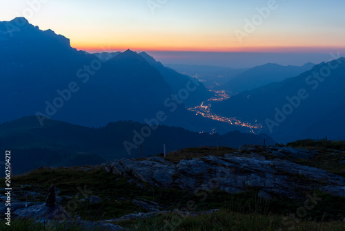 Fotografie Beautiful view of mountain peaks during twilight, with a winding valley of glowing city lights nestled between slopes under a vibrant sunrise sky