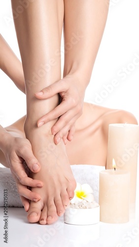 Woman's hand touching a foot, with candles, towel, and flower arranged nearby on a white surface against white background