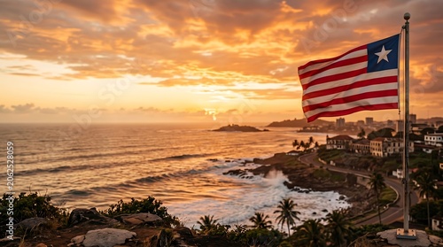 Liberian flag waving over Monrovia coastal city landscape during golden hour sunset with ocean waves