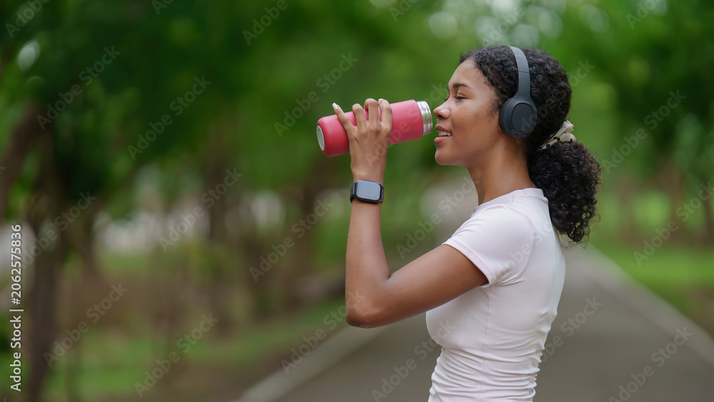 Obraz premium Black woman drinking water from reusable bottle on park path for hydration and wellness
