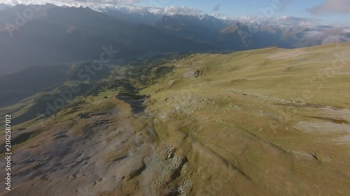 Smooth dive FPV drone flight over open alpine meadows and winding mountain trails, revealing a vast valley, distant snowy peaks, soft clouds, and dramatic high altitude scenery in cinematic motion