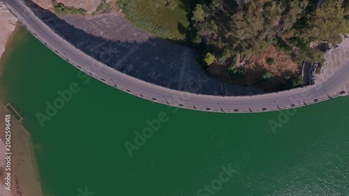 Drone footage spinning directly overhead El Chorro's curved dam crest, abstracting its sweeping arc against a vast jade reservoir, shadowed stone face, and pine-topped embankment.