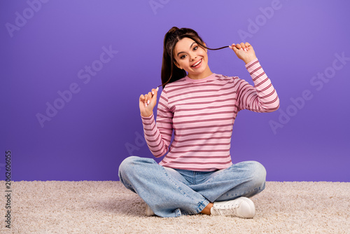 Young woman smiling and playful while sitting cross-legged on carpet against a purple background wearing striped pink top and jeans